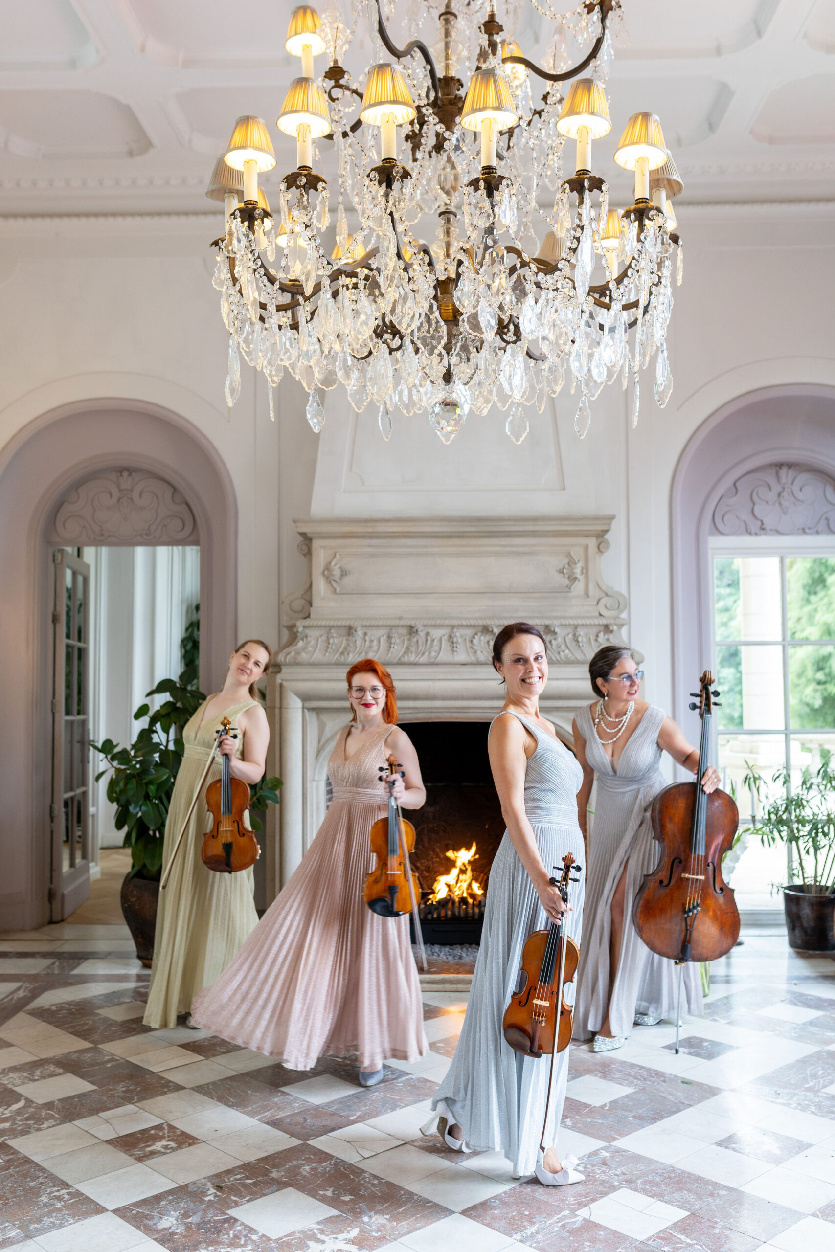 Vier musici in pastelkleurige avondjurken met instrumenten bij een open haard onder een kristallen kroonluchter